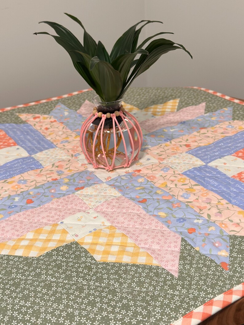 Pu&ograve; includere: Un vaso di vetro trasparente con una gabbia decorativa rosa contiene piante verdi. Il vaso &egrave; posto su una colorata trapunta patchwork con motivi floreali e geometrici nei toni del rosa, blu, giallo e verde.