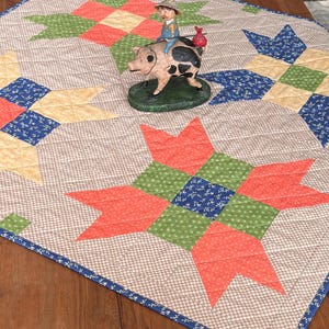 May include: A quilted table runner with a patchwork star design in shades of coral, green, blue, and yellow. The runner has a brown and white checkered pattern and a blue floral border. A figurine of a person riding a pig is on the runner.