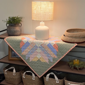 Pu&ograve; includere: Una mensola decorativa con una lampada con paralume in lino, una trapunta fantasia, cesti intrecciati e un vaso con verde. La trapunta presenta un motivo a stella in colori pastello. I cesti hanno manici bianchi.