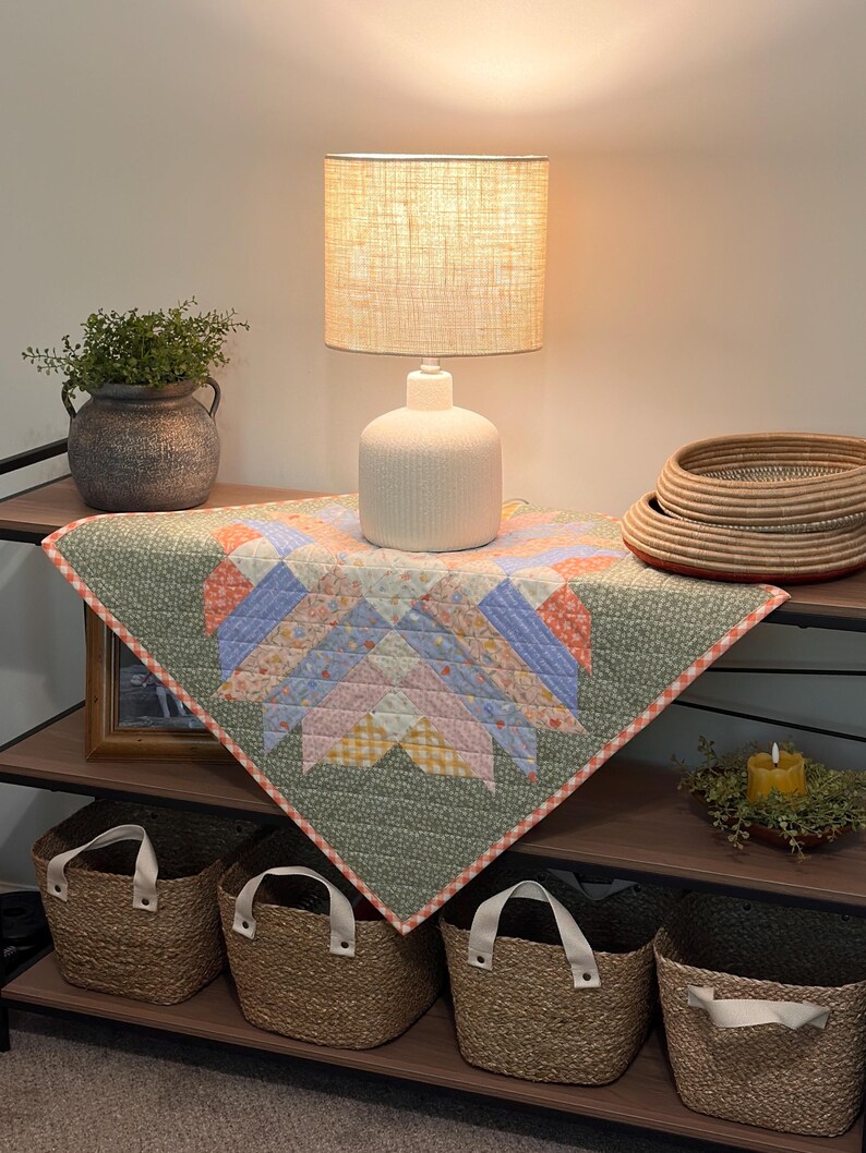 Pu&ograve; includere: Una mensola decorativa con una lampada con paralume in lino, una trapunta fantasia, cesti intrecciati e un vaso con verde. La trapunta presenta un motivo a stella in colori pastello. I cesti hanno manici bianchi.