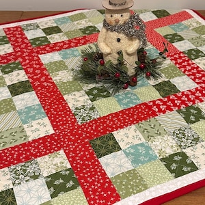 May include: A festive Christmas table runner with a patchwork design in red, green, and white. A snowman figurine with a top hat and scarf sits atop the runner, surrounded by a wreath with red berries.