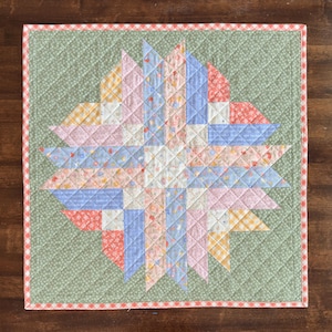 May include: A handmade, square quilt with a starburst design. The quilt features a central star pattern in pastel pink, blue, yellow, and white fabrics. The star is set against a green background with a diamond pattern, and a red and white gingham border.