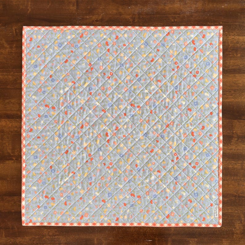 Pu&ograve; includere: Una trapunta quadrata azzurra con un motivo floreale rosso, giallo e blu. La trapunta ha un motivo a rombi e un bordo a quadretti rossi e bianchi. La trapunta misura circa 76 cm quadrati.