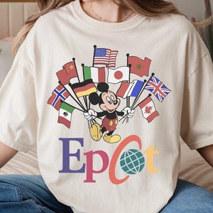 May include: Cream-colored t-shirt featuring a graphic of Mickey Mouse holding flags from various countries. The word "Epcot" is printed in colorful letters below the graphic, with a globe icon. The shirt is a casual style.