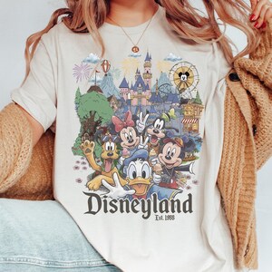 Puede incluir: Una camiseta de color crema con un gr&aacute;fico colorido de personajes de Disney que incluye a Mickey Mouse, Minnie Mouse, el Pato Donald, Goofy y Pluto frente a un castillo, con el texto Disneyland Est. 1955 impreso debajo.