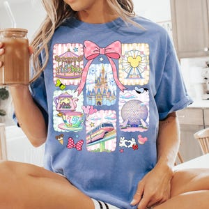 May include: A blue t-shirt featuring a colorful graphic of various theme park icons, including a carousel, castle, and Ferris wheel. The design is framed by a pink bow and decorative borders. The shirt is made of a soft material.