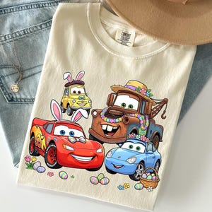 May include: Cream-colored t-shirt featuring cartoon cars decorated for Easter. The graphic includes a red race car, a blue sports car, a brown tow truck, and a yellow car, all with Easter bunny ears and floral decorations. Easter eggs and flowers are scattered around the cars.