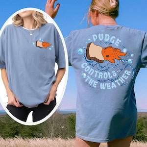 May include: A light blue t-shirt with a graphic of a cartoon fish on a piece of bread, with the text "Pudge Controls the Weather". The shirt has a relaxed fit and short sleeves. The design is on the front and back of the shirt.