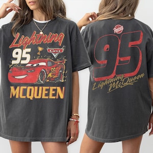 May include: Dark gray t-shirt featuring a graphic of Lightning McQueen from the movie Cars. The front of the shirt displays "Lightning 95 McQueen" with a red race car. The back has a large "95" and "Lightning McQueen" in red.