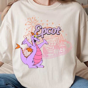 May include: Cream-colored t-shirt featuring a purple dragon character with orange wings and a pink belly. The word "Epcot" is written above the dragon in a stylized font, with a pink outline of a theme park in the background.
