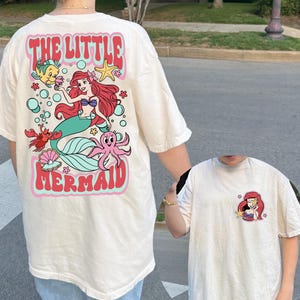 K&ouml;nnte beinhalten: Eine wei&szlig;e Keramik-Tasse mit schwarzem Henkel und dem Text "But First, Coffee" in fetter, schwarzer Schrift. Die Tasse steht vor einem verschwommenen Hintergrund, der eine gem&uuml;tliche Umgebung suggeriert.