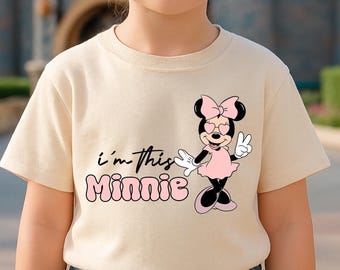May include: A beige t-shirt featuring a Minnie Mouse graphic with heart-shaped sunglasses and a peace sign. The text "i'm this Minnie" is printed in black and pink. The shirt is worn by a person wearing sunglasses.