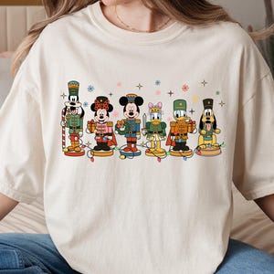 May include: Cream-colored t-shirt featuring a festive graphic of Disney characters dressed as nutcrackers. The design includes Mickey Mouse, Minnie Mouse, Donald Duck, and Goofy, with colorful details and holiday lights. A fun and whimsical design for Disney fans.
