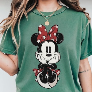 Puede incluir: Camiseta verde con un gr&aacute;fico de Minnie Mouse. El gr&aacute;fico muestra a Minnie Mouse con un lazo rojo de lunares y un dise&ntilde;o en blanco y negro. La camiseta tiene cuello redondo y mangas cortas.