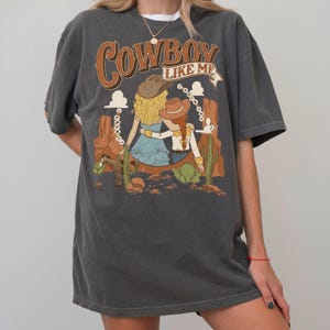 May include: A charcoal grey short-sleeve t-shirt featuring a graphic print of two figures in cowboy hats standing in a desert landscape with cacti and rock formations. The text COWBOY LIKE ME is printed in large brown letters above the figures.