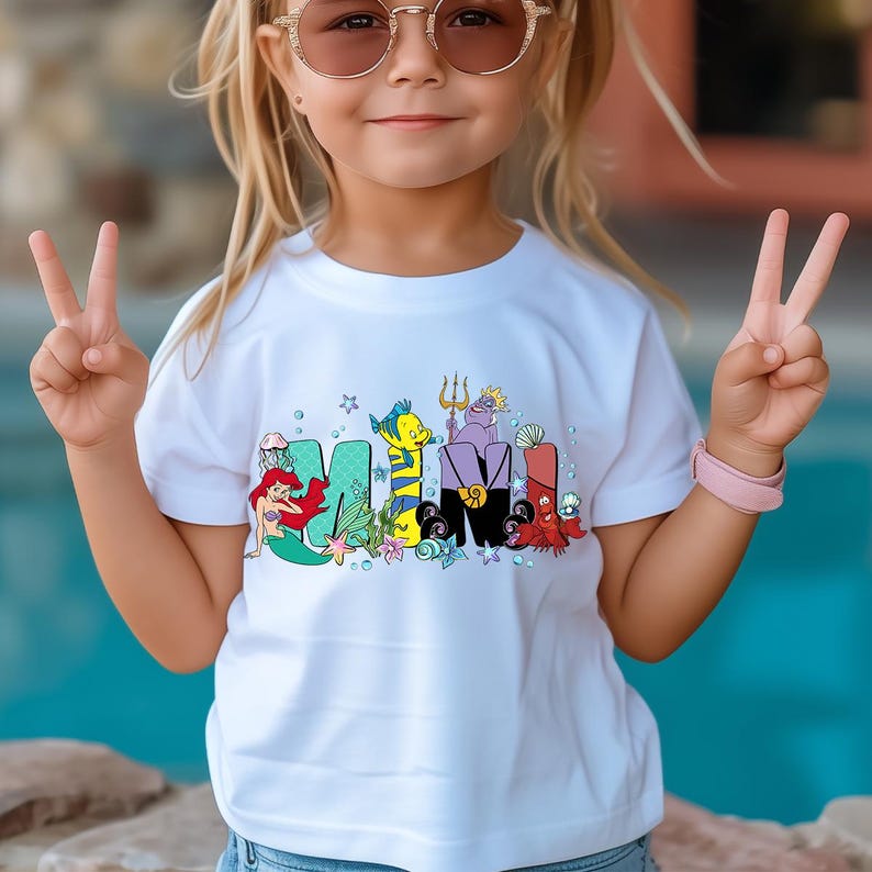 Puede incluir: Camiseta blanca con la palabra "MAMI" en letras de dibujos animados coloridas, con personajes de una pel&iacute;cula de animaci&oacute;n popular. El dise&ntilde;o incluye una sirena, un pez y otras criaturas marinas. El ni&ntilde;o lleva gafas de sol y una pulsera rosa.