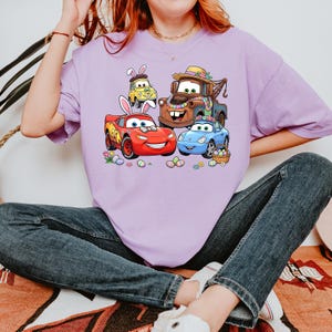 May include: A light purple t-shirt featuring a colorful graphic of animated cars decorated for Easter. The cars include a red race car, a blue car, a brown tow truck, and a yellow car, all with Easter bunny ears and surrounded by Easter eggs.