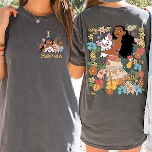 May include: Dark gray t-shirt featuring a colorful graphic of a cartoon character with a pig and flowers on the back. The front of the shirt has a smaller graphic with the name "SOPHIA" in gold lettering. The shirt is made of a soft material.
