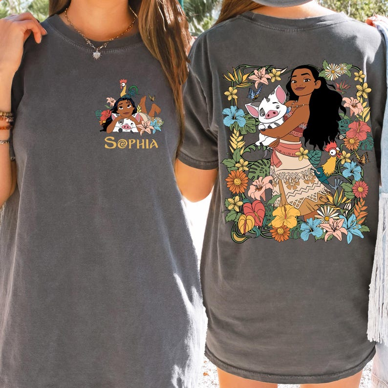 May include: Dark gray t-shirt featuring a colorful graphic of a cartoon character with a pig and flowers on the back. The front of the shirt has a smaller graphic with the name "SOPHIA" in gold lettering. The shirt is made of a soft material.
