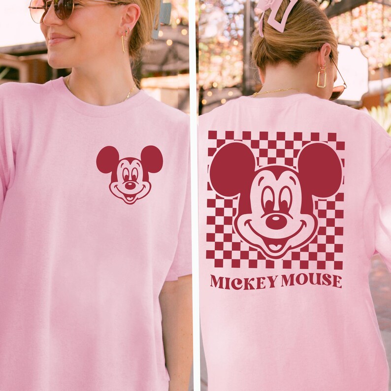 May include: A light pink t-shirt featuring a maroon Mickey Mouse graphic on the front and back. The back design includes a checkered pattern and the words "Mickey Mouse".