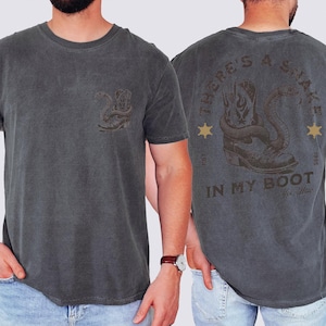 May include: A charcoal gray t-shirt featuring a small graphic on the front left chest of a cowboy boot with a snake. The back displays a large graphic of a cowboy boot with a snake, the text "THERE'S A SNAKE IN MY BOOT", "EST. 1995", and "Yee-Haw".