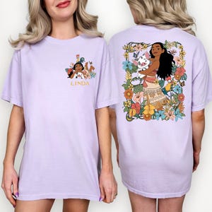 May include: A light purple t-shirt featuring a colorful graphic of a cartoon character on the front and back. The front graphic includes the name "LINDA" in gold. The back graphic shows a character with a pig and surrounded by flowers.