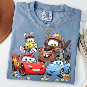 May include: A blue Comfort Colors t-shirt featuring a graphic of animated cars with Easter bunny ears and floral decorations. The cars include a yellow taxi, a red race car, a brown tow truck, and a blue sports car. The shirt also has a gold necklace.