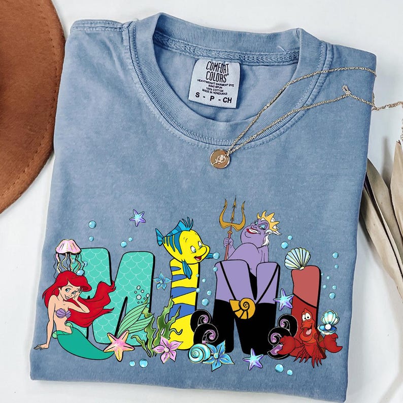 Puede incluir: Una camiseta azul Comfort Colors con la palabra "MOM" y personajes de La Sirenita. La "M" es verde con una sirena, la "O" es amarilla con un pez y la "M" es morada con &Uacute;rsula y Sebasti&aacute;n.