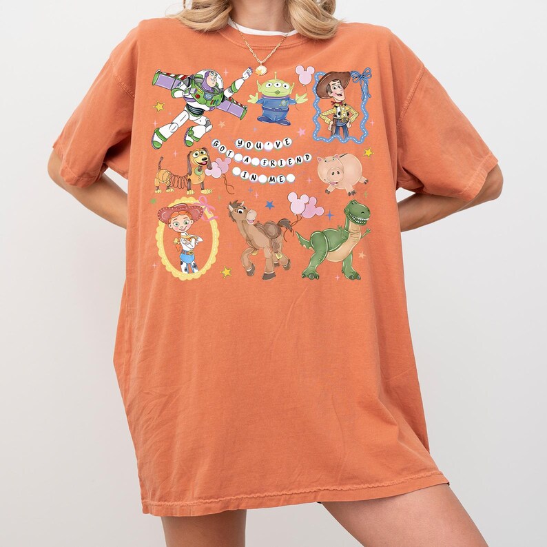 May include: An orange t-shirt featuring a colorful graphic of characters from the animated film Toy Story. The design includes Buzz Lightyear, Woody, and other characters, with the text "You've Got a Friend in Me."