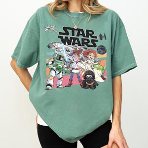 Puede incluir: Camiseta verde salvia con un gr&aacute;fico de Star Wars con personajes de dibujos animados y el logotipo de Star Wars. El dise&ntilde;o incluye personajes como Darth Vader, Woody y Buzz Lightyear. La camiseta tiene un corte holgado.
