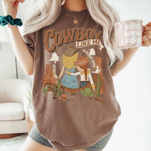 May include: A brown short-sleeve t-shirt featuring a graphic print with the text "COWBOY LIKE ME" and "I KNEW YOU". The illustration depicts two figures in cowboy hats, a horse, and cacti set against a desert landscape with rock formations.