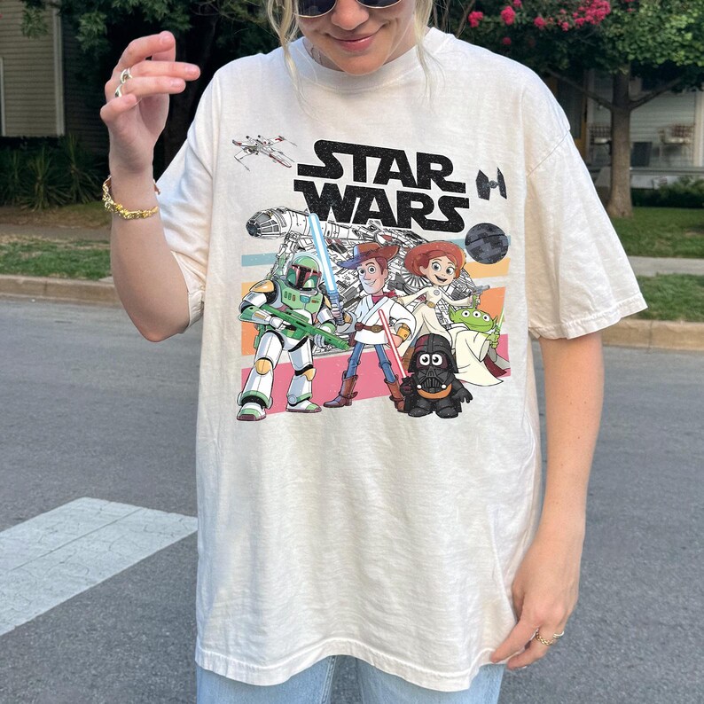 Puede incluir: Camiseta color crema con un gr&aacute;fico de personajes de Star Wars al estilo de dibujos animados. El dise&ntilde;o incluye el logotipo de Star Wars e im&aacute;genes de personajes como Darth Vader, Woody y Buzz Lightyear. La camiseta tiene mangas cortas y un corte holgado.