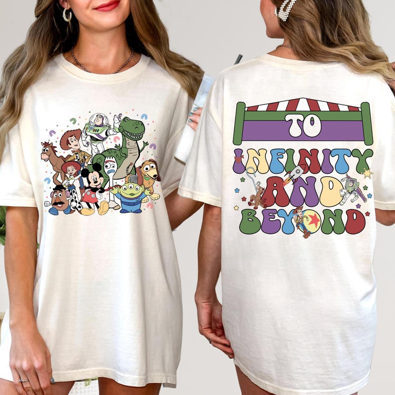 May include: Cream-colored t-shirt featuring a colorful graphic of Toy Story characters on the front. The back of the shirt has the phrase "TO INFINITY AND BEYOND" in a playful font, with a rocket ship and other Toy Story elements.