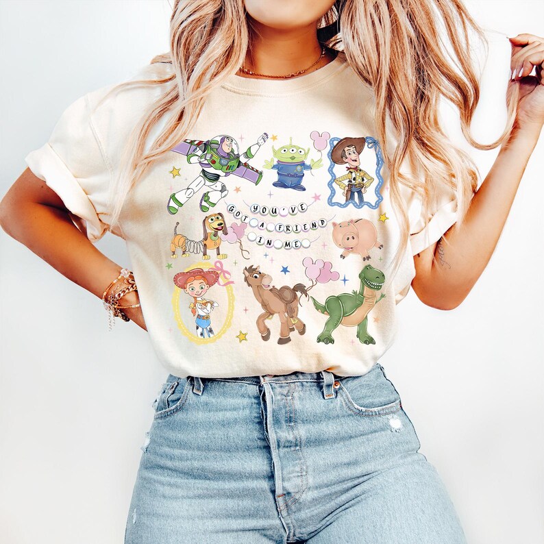May include: Cream-colored t-shirt featuring a colorful graphic of characters from the animated movie Toy Story. The design includes Buzz Lightyear, Woody, the Little Green Men, and other characters, with the text "You've Got a Friend in Me."