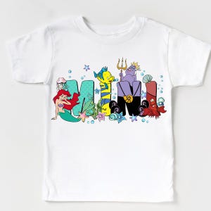 Puede incluir: Camiseta blanca con la palabra "MIMI" en letras de dibujos animados coloridas. Las letras est&aacute;n decoradas con personajes y elementos de una popular pel&iacute;cula de animaci&oacute;n, incluyendo una sirena, un pez y criaturas marinas. La camiseta est&aacute; dise&ntilde;ada para ni&ntilde;os.