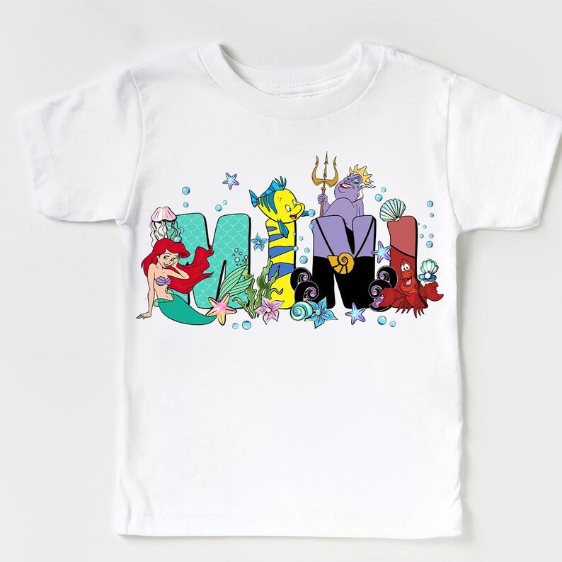 Puede incluir: Camiseta blanca con la palabra "MIMI" en letras de dibujos animados coloridas. Las letras est&aacute;n decoradas con personajes y elementos de una popular pel&iacute;cula de animaci&oacute;n, incluyendo una sirena, un pez y criaturas marinas. La camiseta est&aacute; dise&ntilde;ada para ni&ntilde;os.