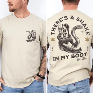 May include: A beige short-sleeve t-shirt featuring a small black graphic of a snake coiled around a cowboy boot on the front left chest. The back displays a large version of the same graphic with the text THERE'S A SNAKE IN MY BOOT, EST. 1995, and Yee-Haw.