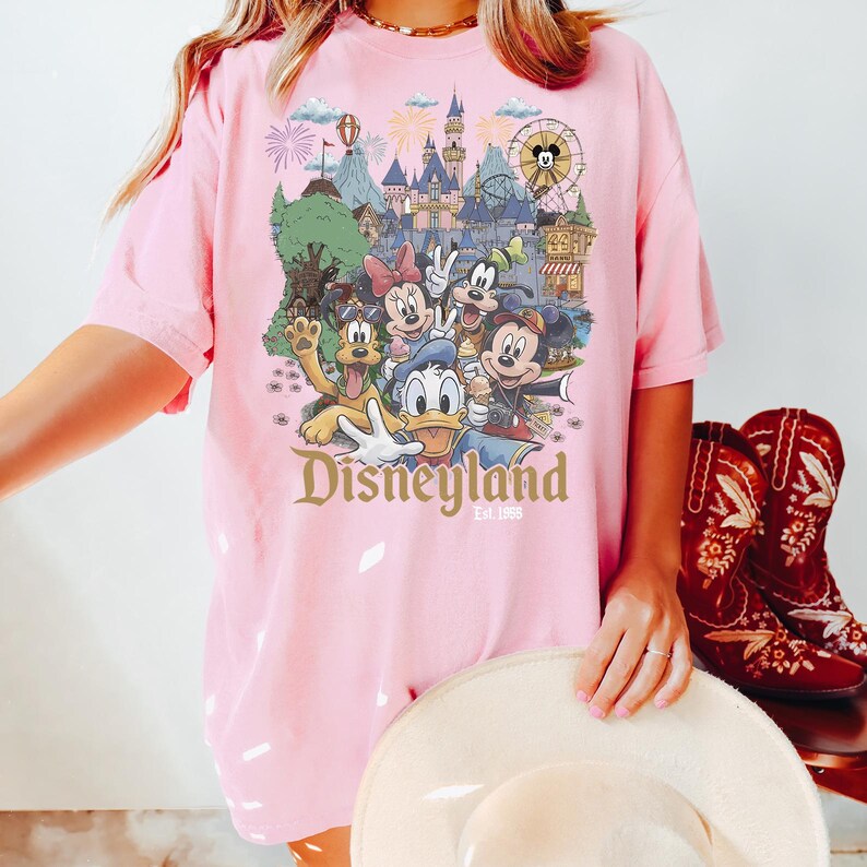 Puede incluir: Una camiseta de manga corta de color rosa claro con un estampado gr&aacute;fico de personajes de Disney, incluidos Mickey Mouse, Minnie Mouse, el Pato Donald, Goofy y Pluto frente a un castillo. El texto Disneyland Est. 1955 aparece debajo.