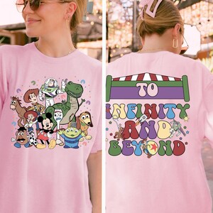 May include: Pink t-shirt featuring a colorful graphic of various cartoon characters, including a dinosaur, a space ranger, and a mouse. The back of the shirt has the text "TO INFINITY AND BEYOND" in a playful font.