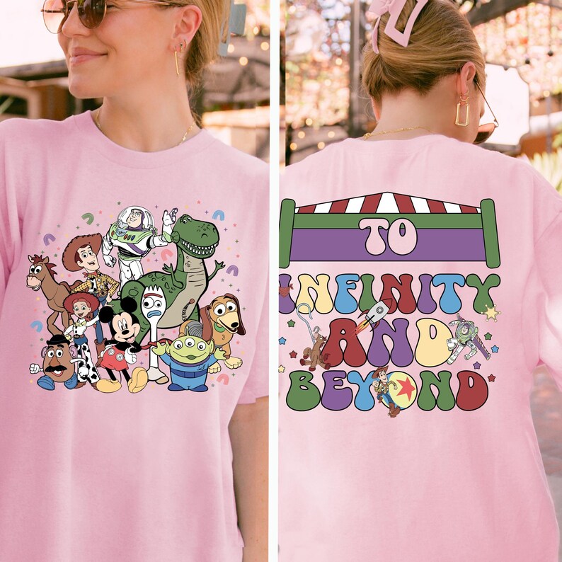 May include: Pink t-shirt featuring a colorful graphic of various cartoon characters, including a dinosaur, a space ranger, and a mouse. The back of the shirt has the text "TO INFINITY AND BEYOND" in a playful font.