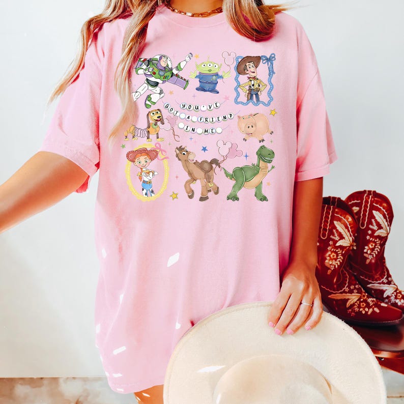 May include: Pink t-shirt featuring a colorful graphic of characters from a popular animated movie. The design includes Buzz Lightyear, Woody, and other characters, along with the phrase "You've Got a Friend in Me."