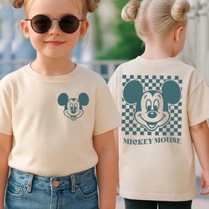 May include: A beige t-shirt featuring a teal Mickey Mouse graphic on the front and back. The back design includes a checkered pattern and the words "Mickey Mouse". The shirt is worn by a young person.