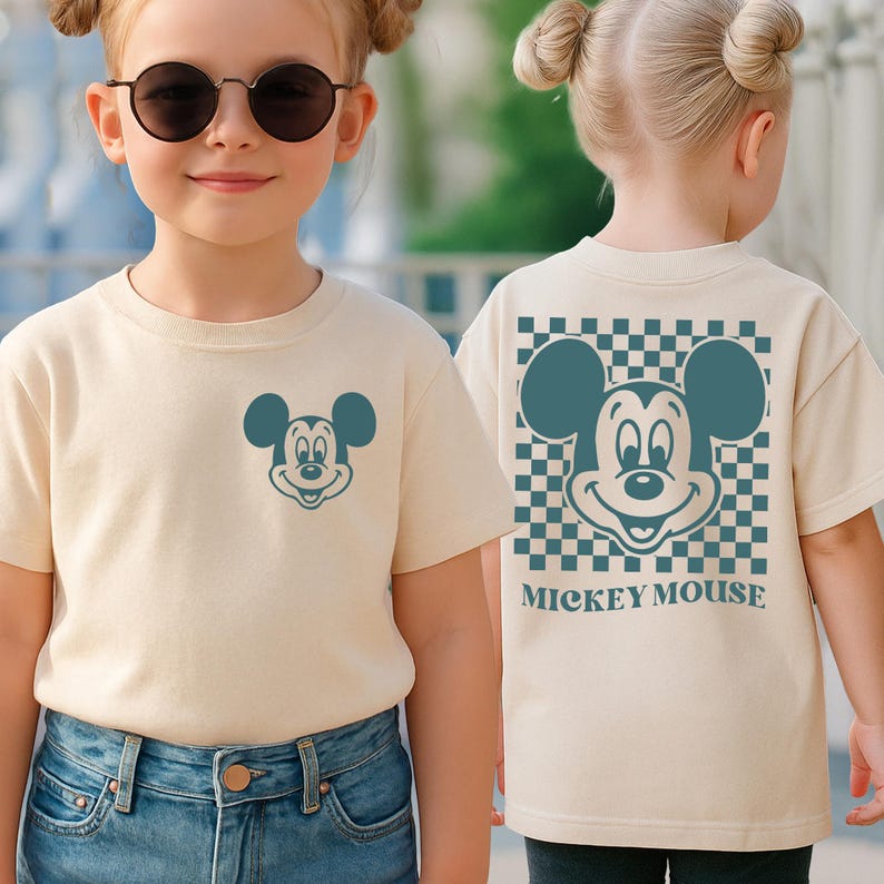 May include: A beige t-shirt featuring a teal Mickey Mouse graphic on the front and back. The back design includes a checkered pattern and the words "Mickey Mouse". The shirt is worn by a young person.