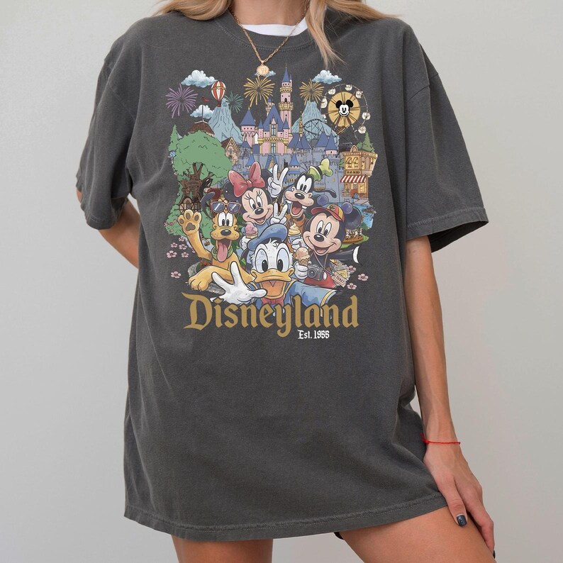 Puede incluir: Una camiseta de manga corta gris carb&oacute;n con un estampado gr&aacute;fico colorido de varios personajes de dibujos animados frente a un castillo. El texto "Disneyland Est. 1955" est&aacute; impreso en letras doradas debajo de los personajes, que incluyen un rat&oacute;n, un pato y un perro.