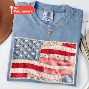 May include: A blue crew neck t-shirt featuring a rectangular patch designed like an American flag. The flag is constructed from various fabric scraps with floral, gingham, and polka dot patterns in red, white, and blue. A gold-toned necklace rests on the shirt.