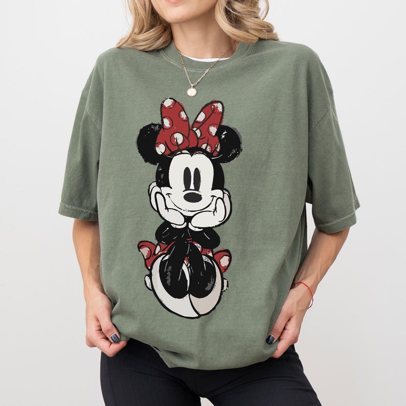 Puede incluir: Camiseta verde oliva con un gran gr&aacute;fico de Minnie Mouse. Minnie se representa sentada, con un lazo rojo de lunares y detalles a juego. La camiseta tiene un corte holgado y mangas cortas.