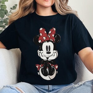 Puede incluir: Camiseta negra con un gr&aacute;fico desgastado de Minnie Mouse. Minnie aparece con un lazo rojo de lunares, orejas negras y una cara sonriente. El gr&aacute;fico est&aacute; centrado en la parte delantera de la camiseta.