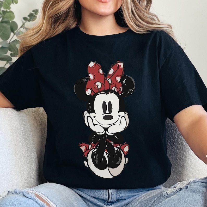 Puede incluir: Camiseta negra con un gr&aacute;fico desgastado de Minnie Mouse. Minnie aparece con un lazo rojo de lunares, orejas negras y una cara sonriente. El gr&aacute;fico est&aacute; centrado en la parte delantera de la camiseta.