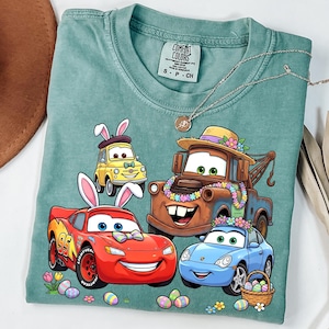 May include: A teal Comfort Colors t-shirt featuring a graphic of animated cars decorated with Easter and spring elements. The cars include Lightning McQueen, Mater, and others, with bunny ears and floral accents. A silver necklace is draped across the shirt.