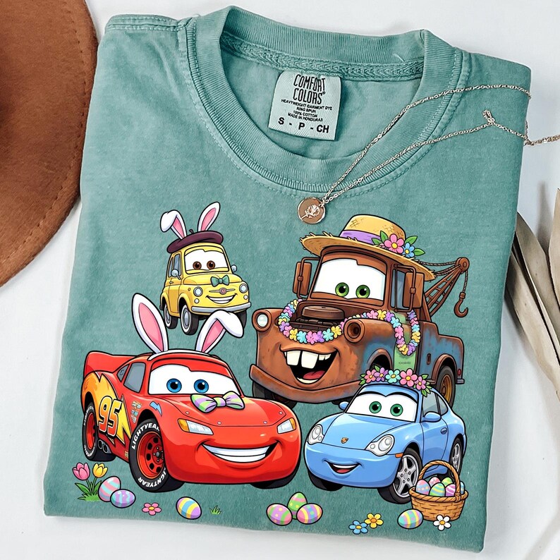 May include: A teal Comfort Colors t-shirt featuring a graphic of animated cars decorated with Easter and spring elements. The cars include Lightning McQueen, Mater, and others, with bunny ears and floral accents. A silver necklace is draped across the shirt.